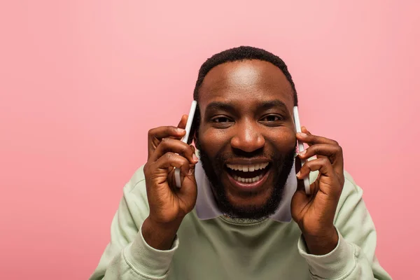 Усміхнений афроамериканський чоловік розмовляє на смартфонах ізольовано на рожевому — стокове фото