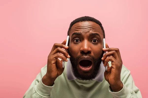 Choqué homme afro-américain parlant sur les smartphones et regardant la caméra isolée sur rose — Photo de stock