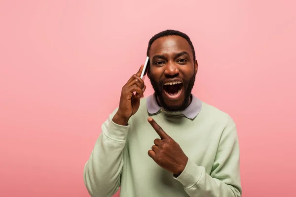 Homem americano africano alegre apontando com o dedo enquanto fala no smartphone isolado no rosa — Fotografia de Stock