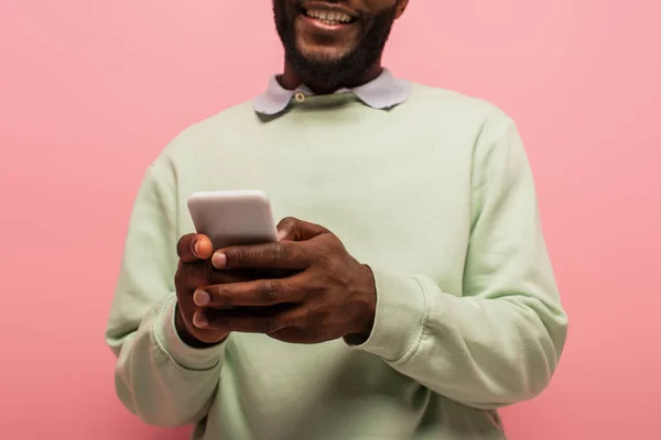 Vista recortada del teléfono inteligente en manos del hombre afroamericano sonriente sobre fondo borroso aislado en rosa - foto de stock