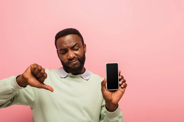 Skeptischer Afroamerikaner hält Handy in der Hand und zeigt Daumen nach unten isoliert auf rosa — Stockfoto