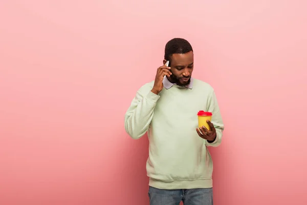 Uomo afroamericano con caffè per andare a parlare sul cellulare su sfondo rosa — Foto stock