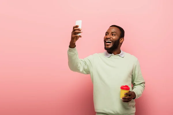 Sonriente hombre afroamericano con taza de papel tomando selfie en el teléfono inteligente sobre fondo rosa - foto de stock