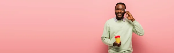 Hombre afroamericano positivo hablando por teléfono móvil y sosteniendo bebida para llevar sobre fondo rosa, pancarta - foto de stock