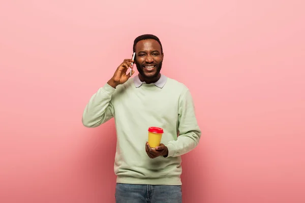 Uomo afroamericano con caffè per andare a parlare al cellulare su sfondo rosa — Foto stock