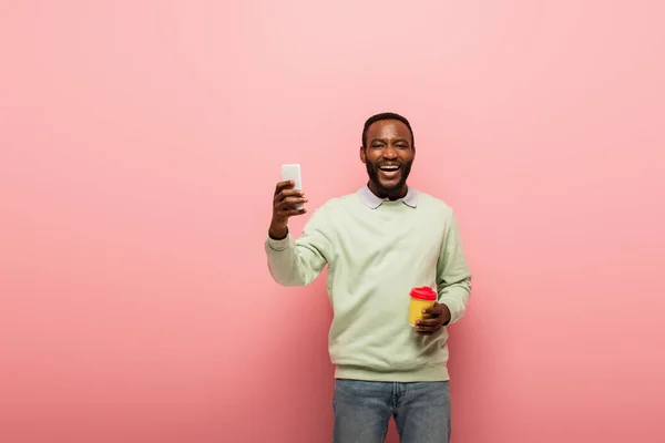 Un homme afro-américain positif tenant son téléphone portable et son café pour aller sur fond rose — Photo de stock
