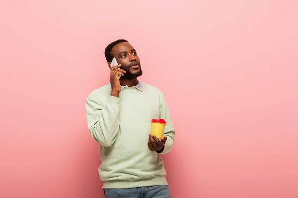 Africano americano homem falando no celular e segurando café para ir no fundo rosa — Fotografia de Stock