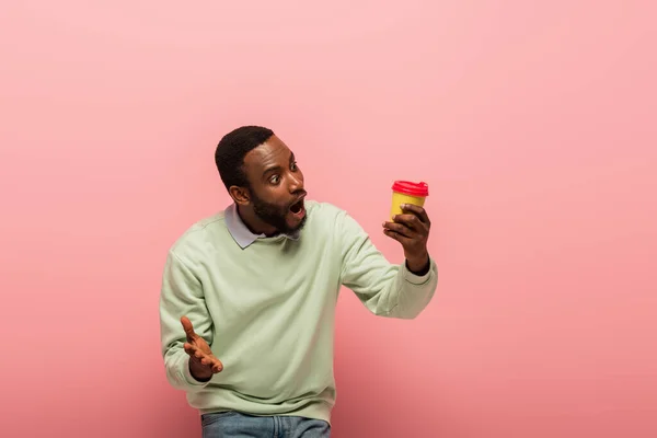 Homem afro-americano chocado olhando para o café para ir em fundo rosa — Fotografia de Stock