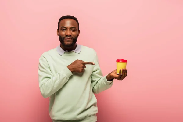 Uomo afroamericano guardando la fotocamera e indicando tazza di carta su sfondo rosa — Foto stock