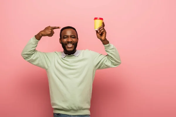 Fröhlicher afrikanisch-amerikanischer Mann hält Coffee to go in der Hand und zeigt mit dem Finger auf rosa Hintergrund — Stockfoto