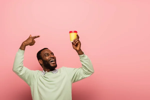 Un homme afro-américain positif pointant du doigt une boisson à emporter sur fond rose — Photo de stock