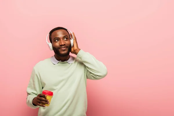Hombre afroamericano con auriculares sosteniendo taza de papel sobre fondo rosa - foto de stock