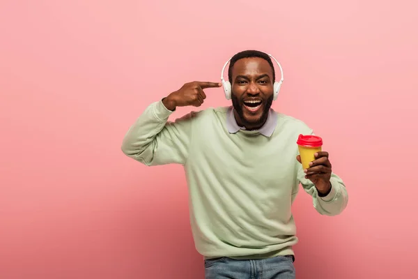 Joyeux homme afro-américain pointant du doigt les écouteurs et tenant du café pour aller sur fond rose — Photo de stock