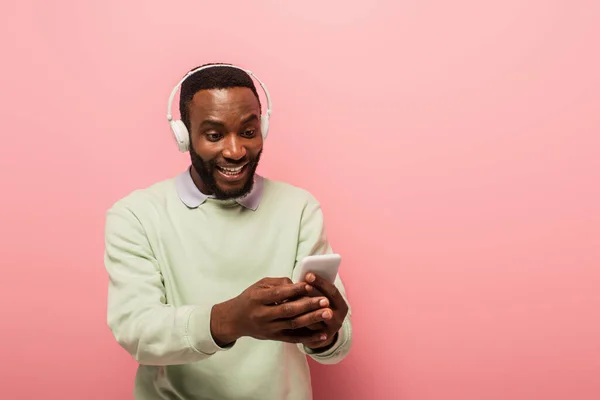 Africano americano em fones de ouvido usando telefone celular e sorrindo em fundo rosa — Fotografia de Stock