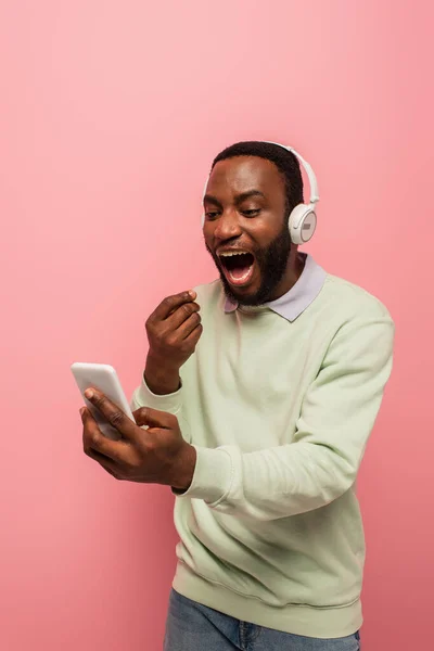 Homem americano africano surpreendente em fones de ouvido usando telefone celular isolado em rosa — Fotografia de Stock