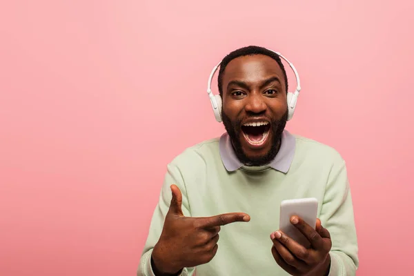 Verblüffter afrikanisch-amerikanischer Mann mit Kopfhörer zeigt auf Smartphone isoliert auf rosa — Stockfoto