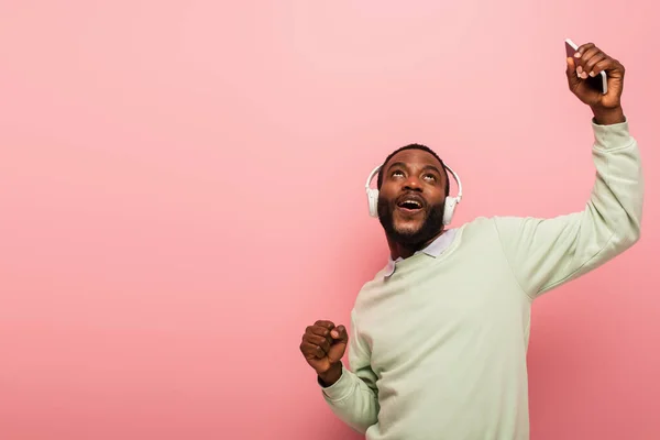 African american man in headphones showing yes gesture and holding mobile phone isolated on pink — Stock Photo