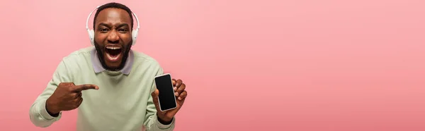 Hombre afroamericano en auriculares apuntando a teléfono inteligente con pantalla en blanco aislado en rosa, bandera - foto de stock