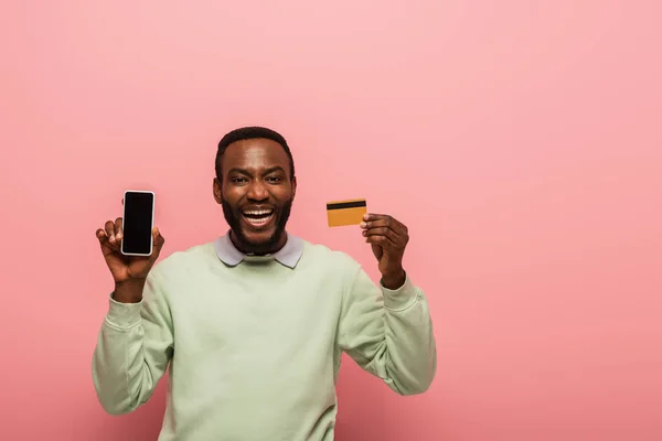 Веселый африканский американец держит мобильный телефон с чистым экраном и кредитной картой на розовом фоне — стоковое фото