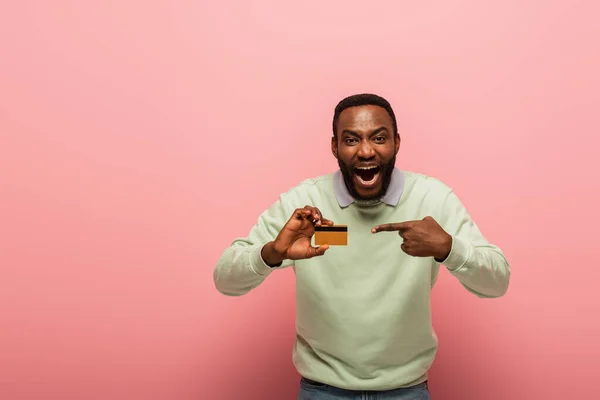 Shocked african american man pointing with finger at credit card on pink background — Stock Photo
