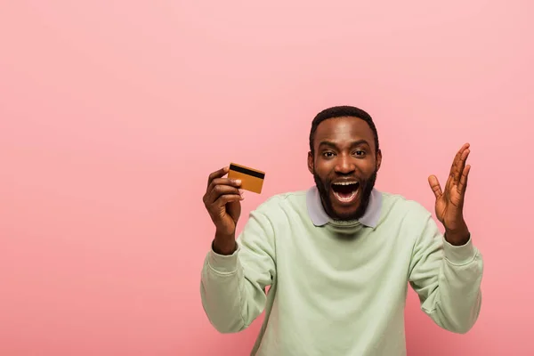 Hombre afroamericano asombrado mirando la cámara mientras que muestra la tarjeta de crédito aislada en rosa - foto de stock