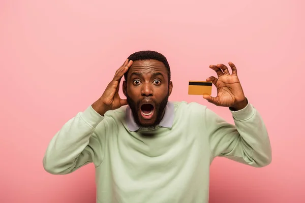 Sorprendido hombre afroamericano tocando la cabeza mientras sostiene la tarjeta de crédito aislado en rosa - foto de stock