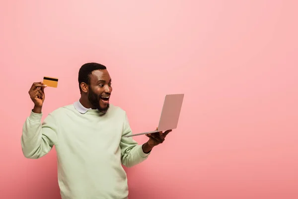 Astonished african american man holding credit card and laptop on pink background — Stock Photo