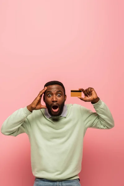 Homme afro-américain ravi tenant carte de crédit et la tête touchante isolé sur rose — Photo de stock