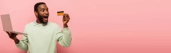 Asombrado hombre afroamericano mirando a la tarjeta de crédito mientras sostiene el ordenador portátil sobre fondo rosa, pancarta - foto de stock