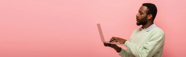 Jeune homme afro-américain tapant sur ordinateur portable isolé sur rose, bannière — Photo de stock