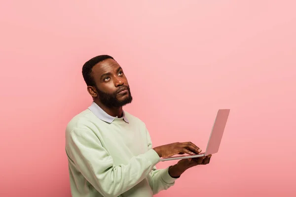 Nachdenklicher afrikanisch-amerikanischer Mann schaut weg, während er Laptop isoliert auf rosa verwendet — Stockfoto
