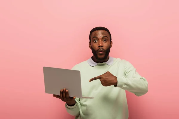 Hombre afroamericano sorprendido apuntando a la computadora portátil en el fondo rosa - foto de stock