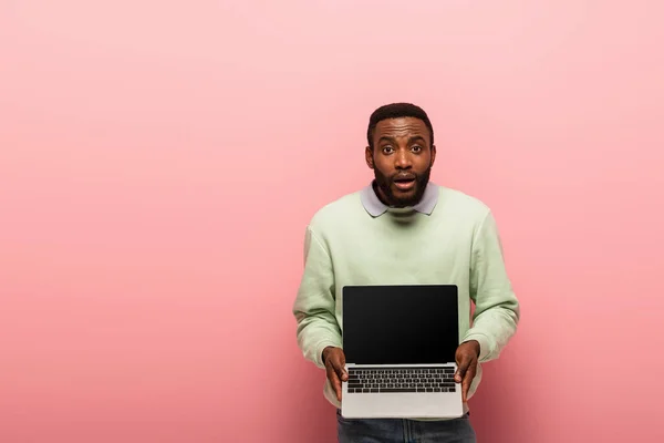 Homme afro-américain choqué montrant ordinateur portable avec écran blanc sur fond rose — Photo de stock