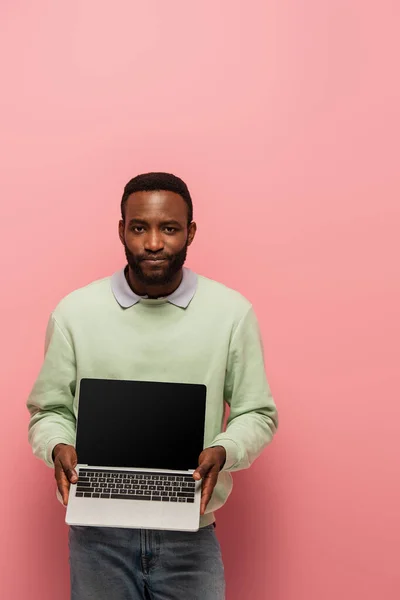 Afrikanisch-amerikanischer Mann blickt in die Kamera, während er Laptop mit leerem Bildschirm auf rosa Hintergrund hält — Stockfoto