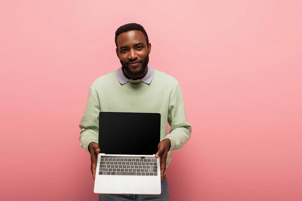 Souriant homme afro-américain tenant ordinateur portable avec écran blanc sur fond rose — Photo de stock