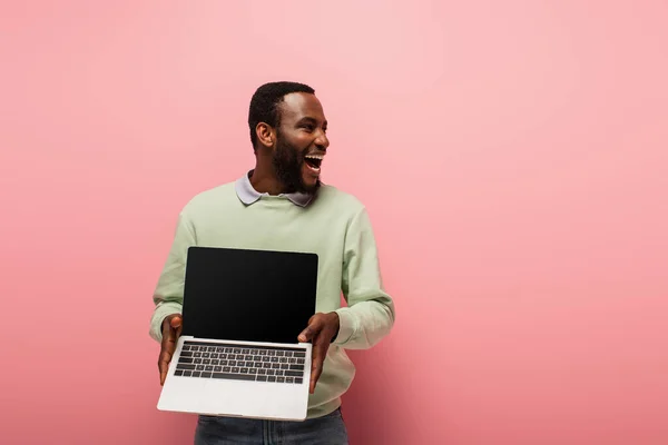 Stupito uomo africano americano guardando lontano e tenendo il computer portatile con schermo bianco su sfondo rosa — Foto stock