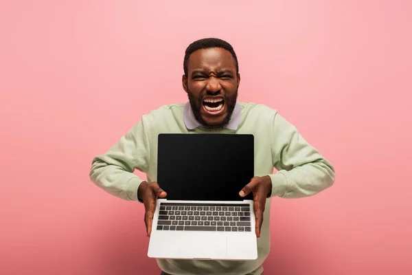 Hombre afroamericano emocionado con la boca abierta que muestra el ordenador portátil con la pantalla en blanco en rosa - foto de stock
