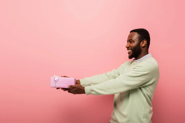 Joyeux homme afro-américain tenant présent dans les mains tendues sur fond rose — Photo de stock