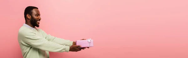 Sorrindo afro-americano homem segurando caixa de presente em mãos estendidas isolado em rosa, banner — Fotografia de Stock