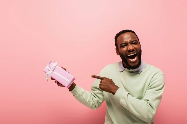 Stupito uomo africano americano che indica scatola regalo isolato su rosa — Foto stock