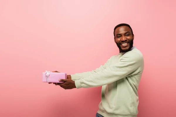 Joyeux homme afro-américain tenant cadeau dans les mains tendues sur fond rose — Photo de stock