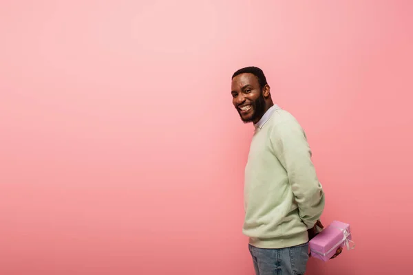 Sourire homme afro-américain tenant boîte-cadeau derrière le dos sur fond rose — Photo de stock