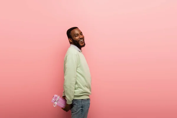 Homme afro-américain joyeux regardant la caméra tout en se cachant présent derrière le dos sur fond rose — Photo de stock