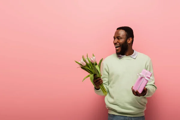 Сміється афроамериканський чоловік з подарунковою коробкою і тюльпанами, дивлячись на рожевий фон — стокове фото