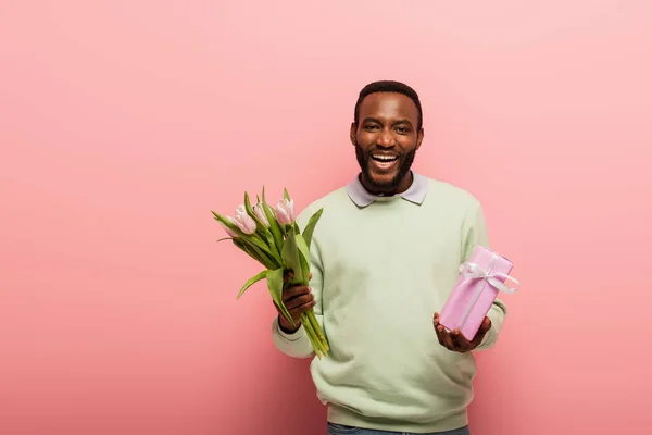 Fröhlicher afrikanisch-amerikanischer Mann mit Geschenkbox und Tulpen lacht in die Kamera auf rosa Hintergrund — Stockfoto