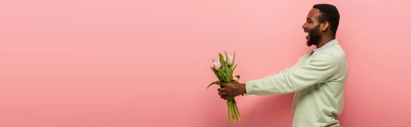 Веселий афроамериканський чоловік тримає свіжі тюльпани в простягнутих руках на рожевому фоні, банер — стокове фото