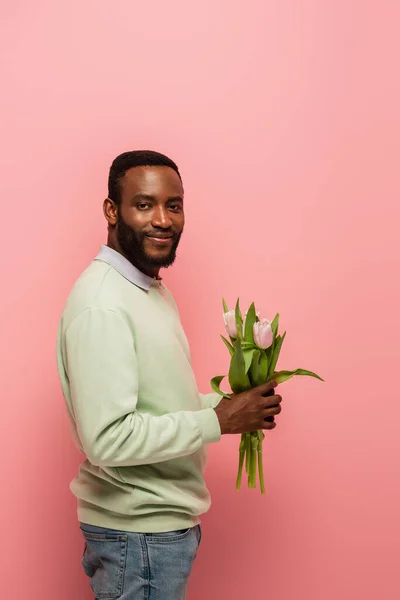 Erfreut afrikanisch-amerikanischer Mann blickt in die Kamera, während er Tulpen isoliert auf rosa hält — Stockfoto