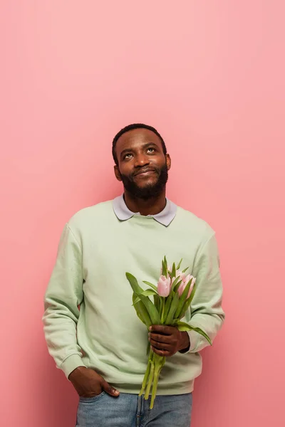 Hombre afroamericano soñador y sonriente sosteniendo tulipanes y mirando hacia arriba sobre fondo rosa - foto de stock