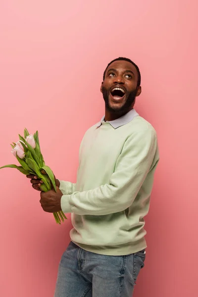 Asombrado hombre afroamericano mirando hacia otro lado mientras sostiene tulipanes sobre fondo rosa - foto de stock