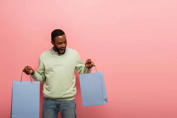 Erstaunt afrikanisch-amerikanischer Mann beim Betrachten von Einkaufstaschen auf rosa Hintergrund — Stockfoto